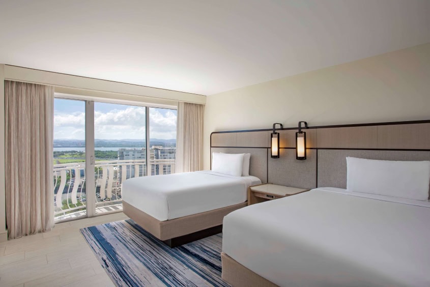 Two beds in a hotel room with a panoramic view of the city skyline through large windows.