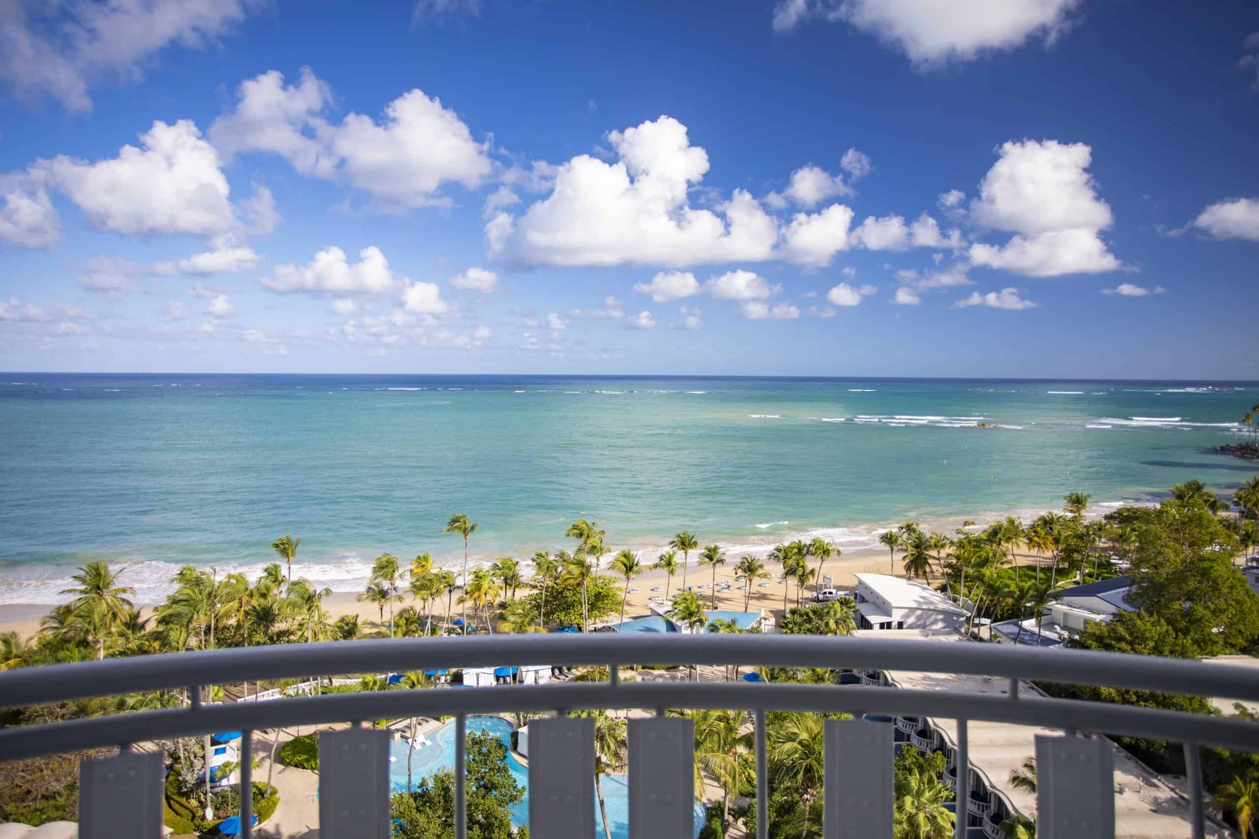 A scenic view of the beach from a hotel balcony, showcasing golden sand and gentle waves under a clear blue sky.