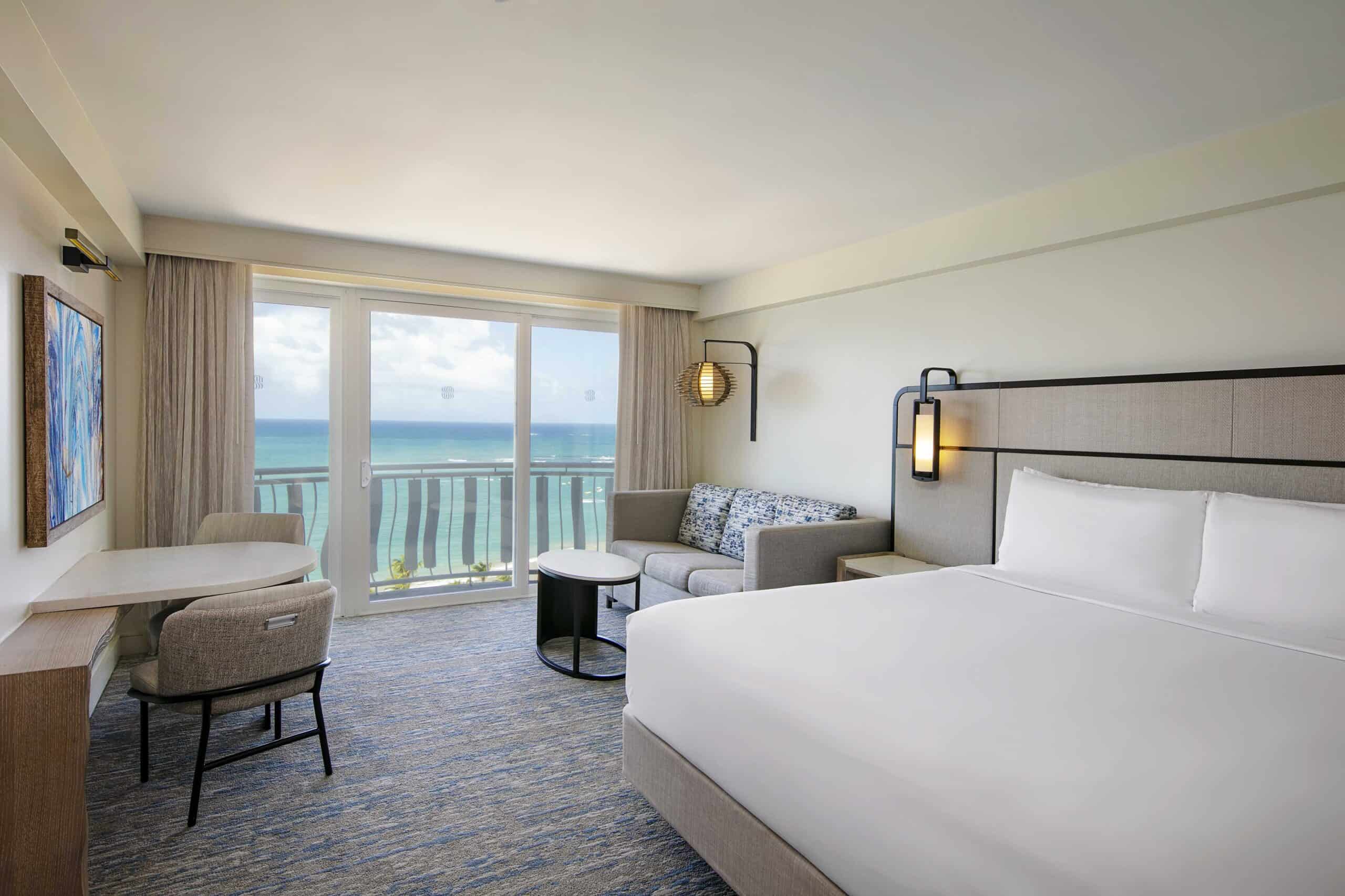 A hotel room featuring a bed, desk, and a large window showcasing a beautiful ocean view.