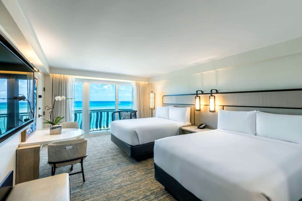Two neatly made beds in a hotel room with a large window showcasing a beautiful ocean view.