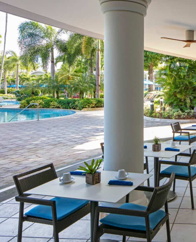 Outdoor dining area featuring tables and chairs adjacent to a swimming pool, surrounded by greenery.