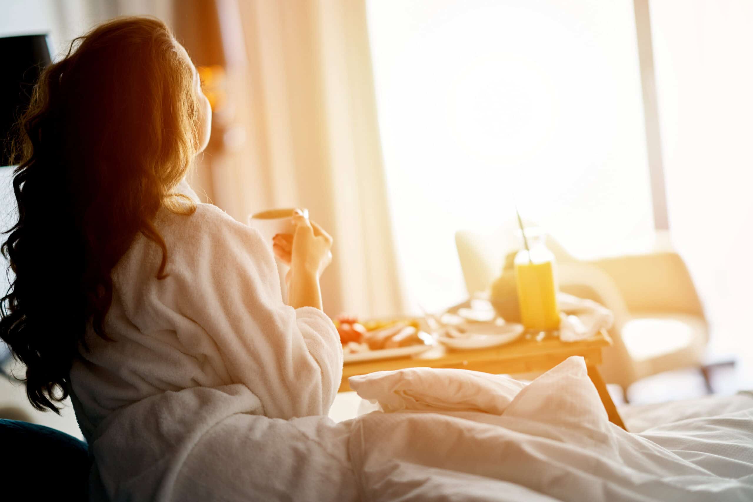A woman in a robe sits in bed, holding a cup of coffee, enjoying a moment of relaxation in the morning light.