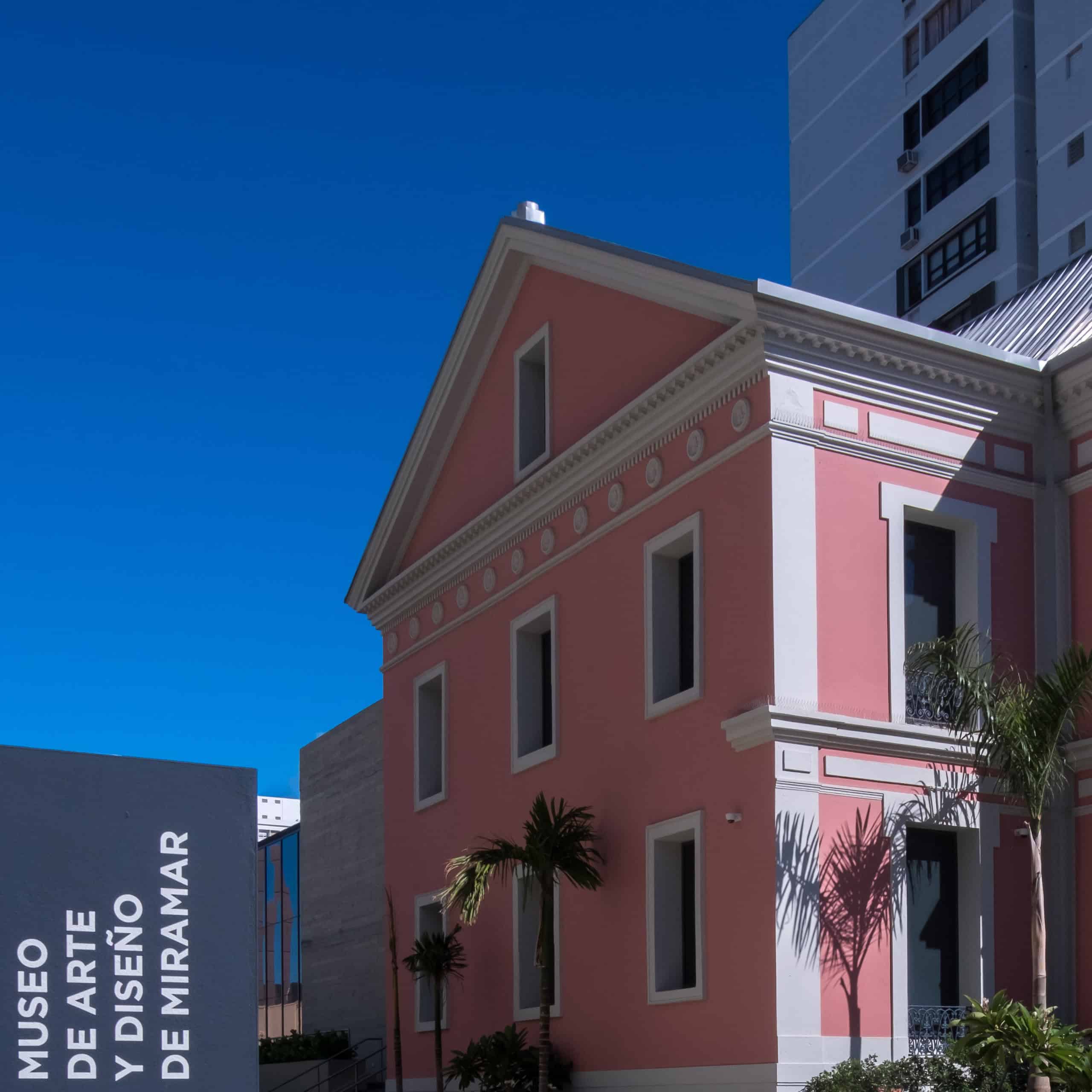 A pink building stands beside a tall palm tree against a clear blue sky.