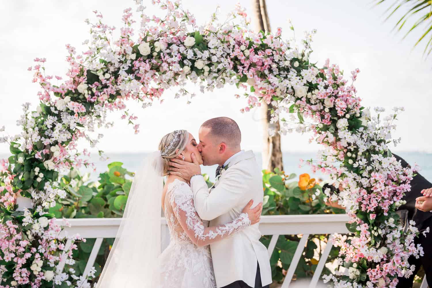 A wedding ceremony taking place on a sandy beach, with an arch and guests seated in the background.