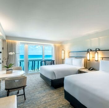 Two neatly made beds in a hotel room with a large window showcasing a beautiful ocean view.
